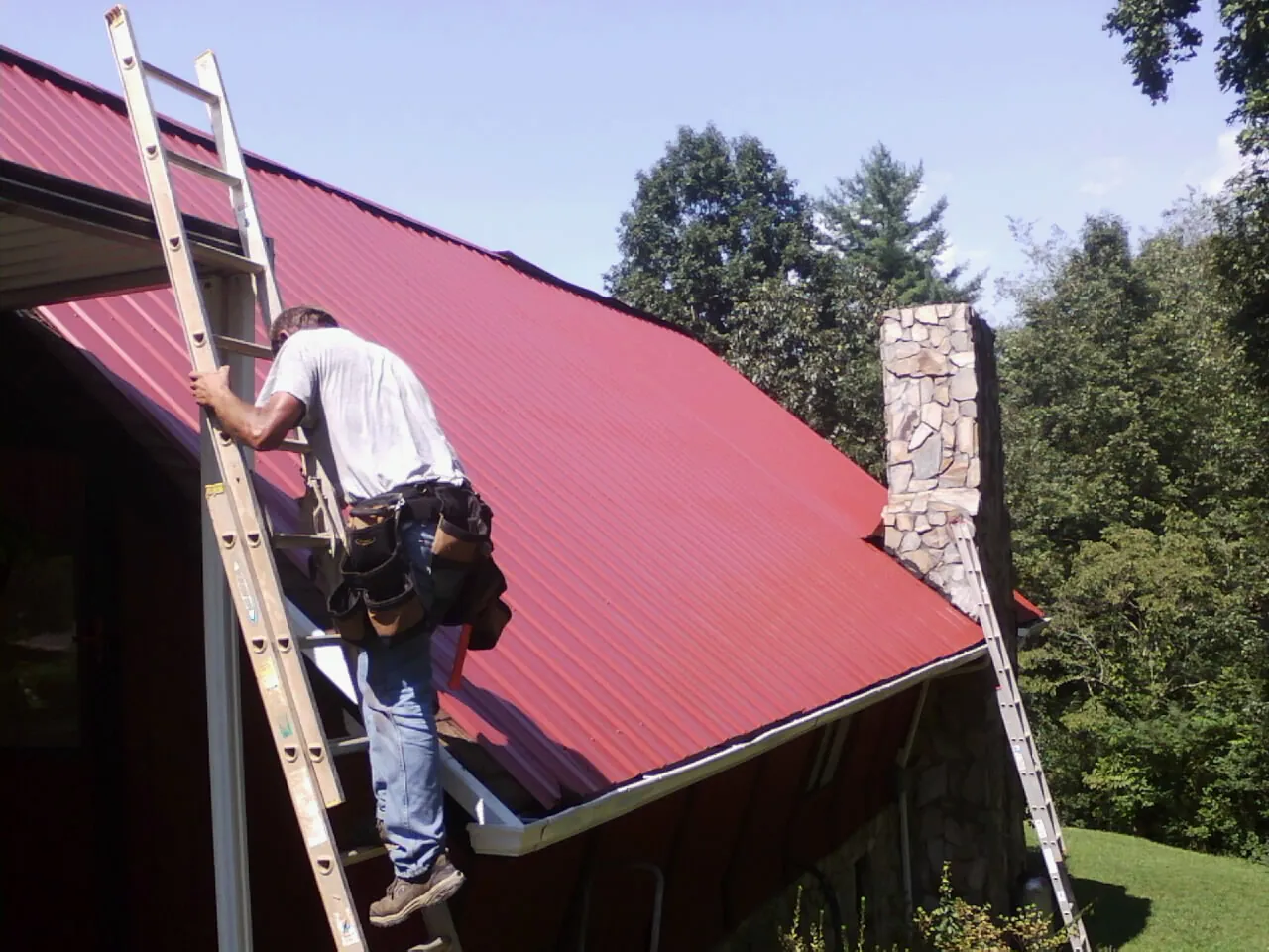 Roofer on a ladder beside a red metal roof and stone chimney — Soffit & Fascia Repair by Roofing Brothers in Holmesville